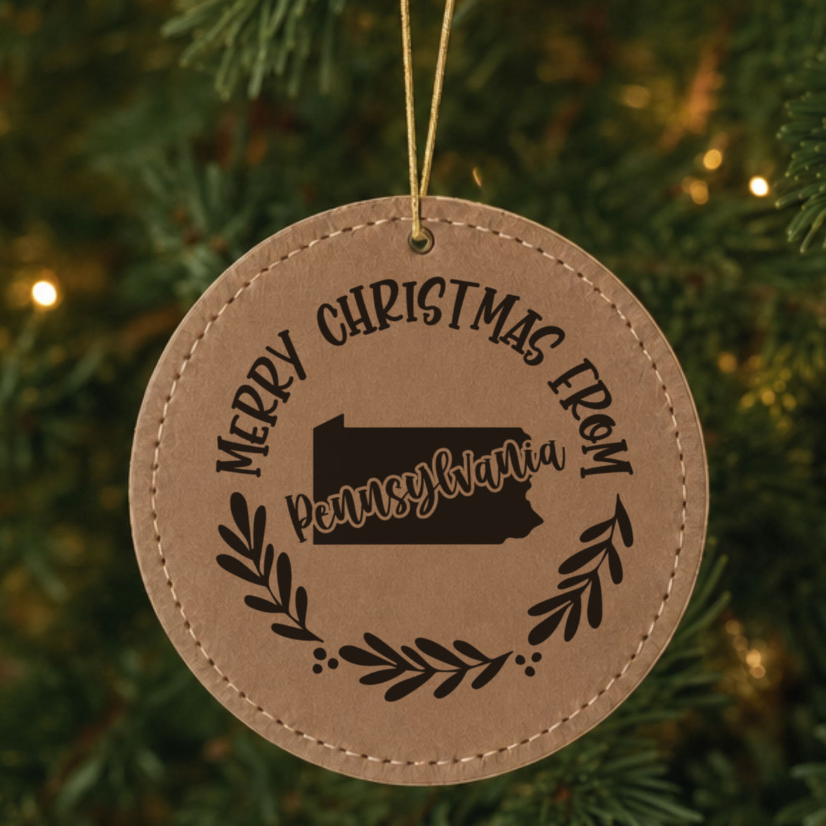Brown leatherette round Christmas ornament engraved Merry Christmas From Pennsylvania, with the state shape in a laurel wreath, hanging on a tree