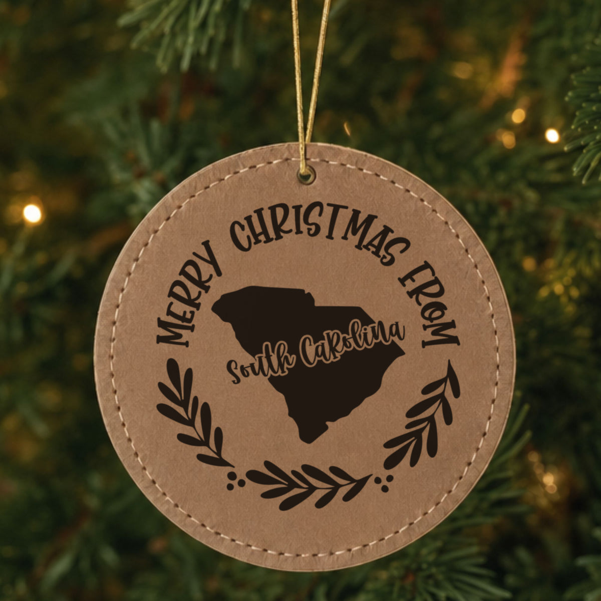 Brown leatherette round Christmas ornament engraved Merry Christmas From South Carolina, with the state shape in a laurel wreath, hanging on a tree