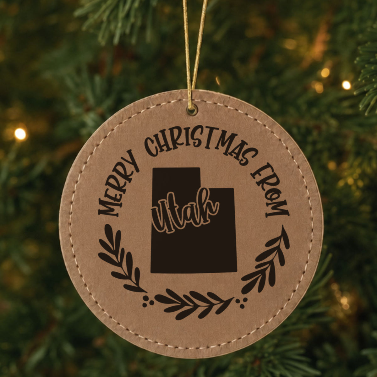 Brown leatherette round Christmas ornament engraved Merry Christmas From Utah, with the state shape in a laurel wreath, hanging on a tree