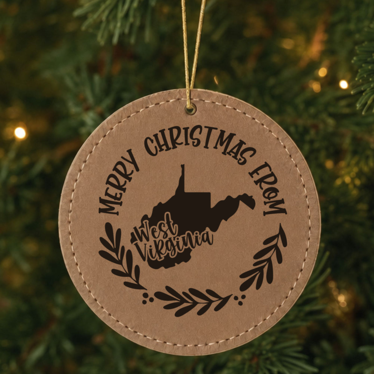 Brown leatherette round Christmas ornament engraved Merry Christmas From West Virginia, with the state shape in a laurel wreath, hanging on a tree