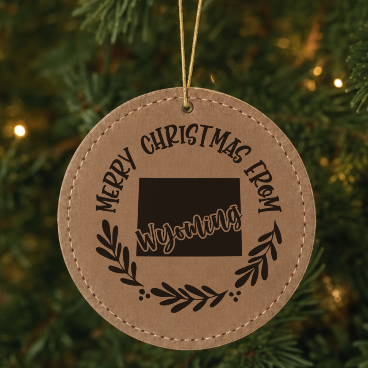 Brown leatherette round Christmas ornament engraved Merry Christmas From Wyoming, with the state shape in a laurel wreath, hanging on a tree