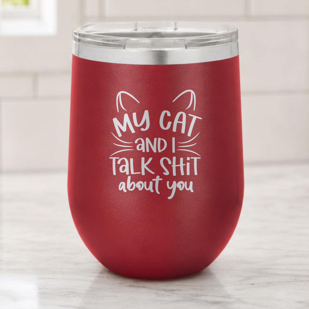 Maroon 12 oz stemless wine tumbler laser engraved with "My Cat and I Talk Sh*t About You", plus cat ears and whiskers, on a kitchen counter