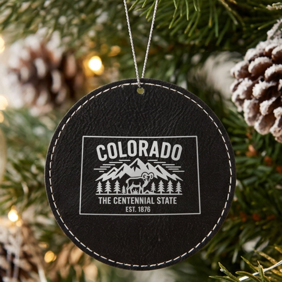 Black leatherette round Christmas ornament laser engraved with a Colorado design of snowcapped peaks, pine forest and a bighorn sheep, hanging on a tree