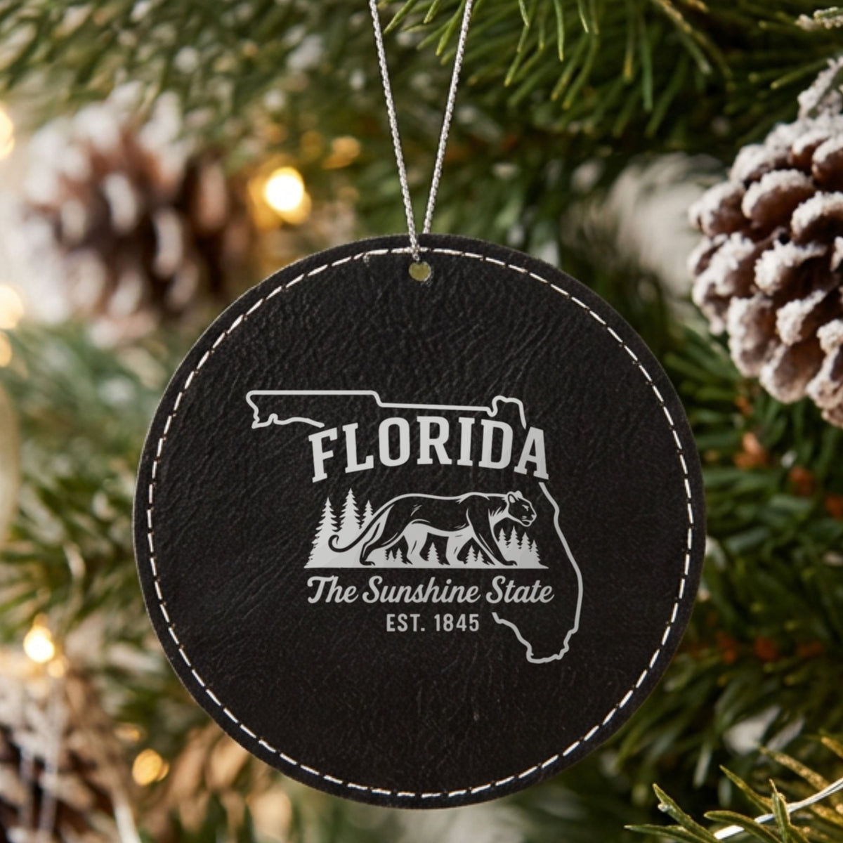 Black leatherette round Christmas ornament laser engraved with a Florida design of a Florida panther and pine forest, hanging on a tree