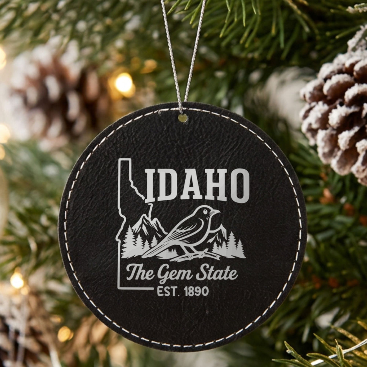 Black leatherette round Christmas ornament laser engraved with an Idaho design of a mountain bluebird, mountains and pines, hanging on a tree