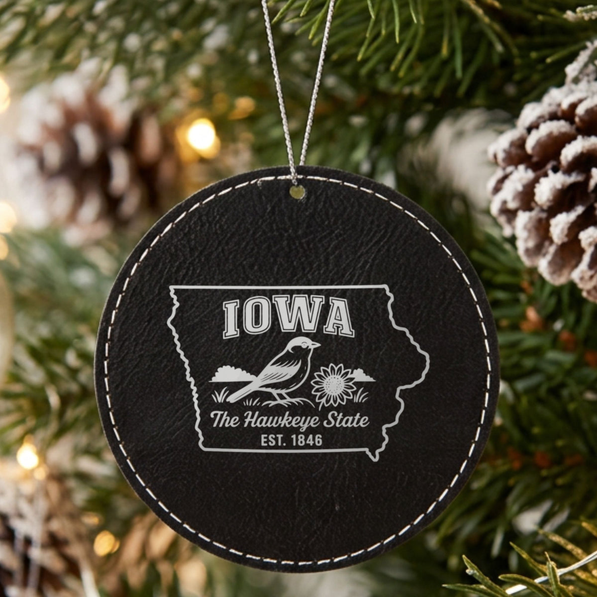 Black leatherette round Christmas ornament laser engraved with an Iowa design of an American goldfinch and a sunflower, hanging on a tree