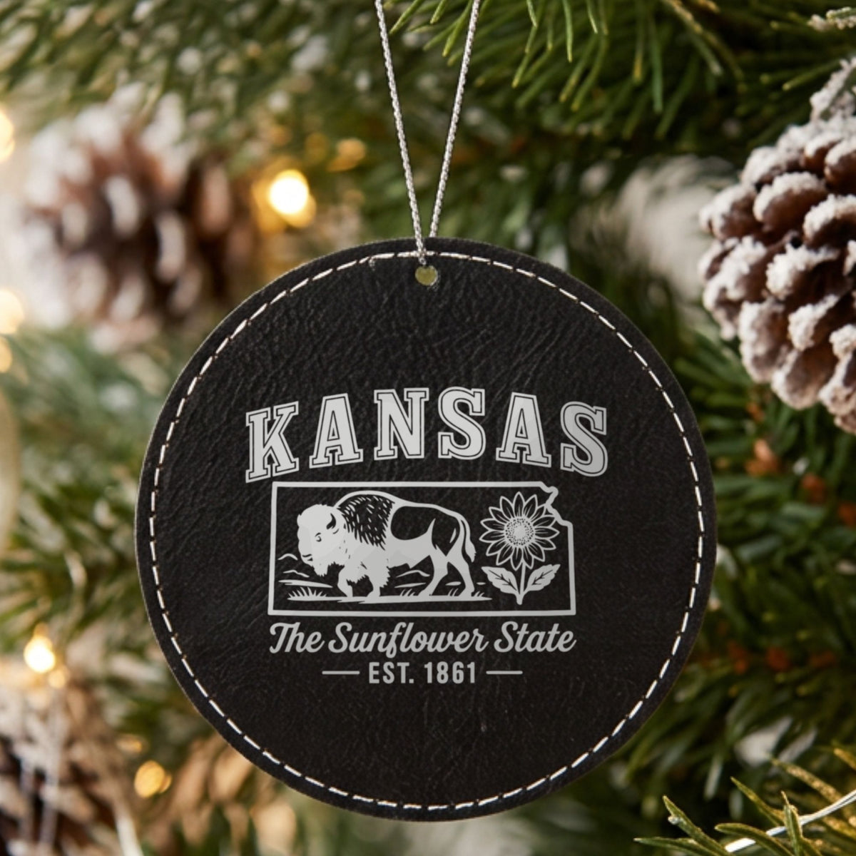 Black leatherette round Christmas ornament laser engraved with a Kansas design of a bison and a sunflower, hanging on a tree