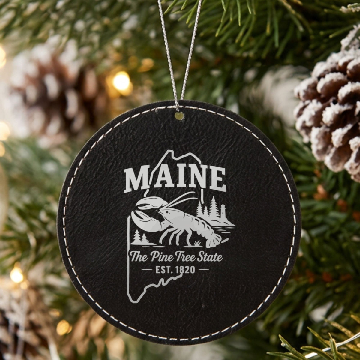 Black leatherette round Christmas ornament laser engraved with a Maine design of a lobster, pines and a lighthouse, hanging on a tree