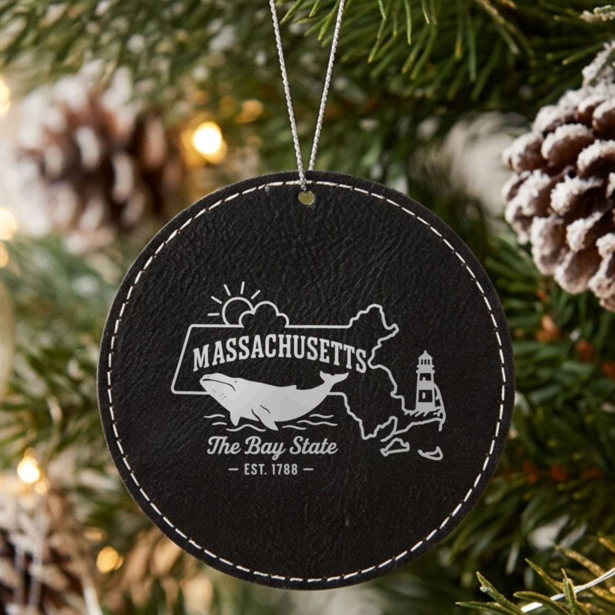 Black leatherette round Christmas ornament laser engraved with a Massachusetts design of a whale, a lighthouse and the sun over Cape Cod and the islands, hanging on a tree