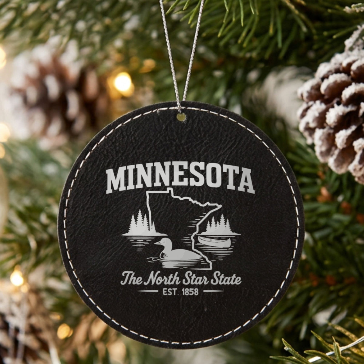 Black leatherette round Christmas ornament laser engraved with a Minnesota design of a loon, a canoe and lakeside pines, hanging on a tree