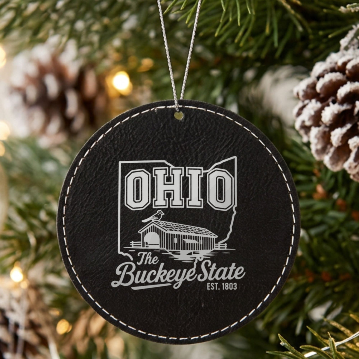 Black leatherette round Christmas ornament laser engraved with an Ohio design of a covered bridge and a cardinal, hanging on a tree