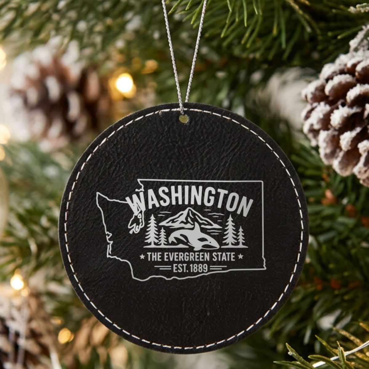 Black leatherette round Christmas ornament laser engraved with a Washington design of an orca, Mount Rainier and evergreens, hanging on a tree