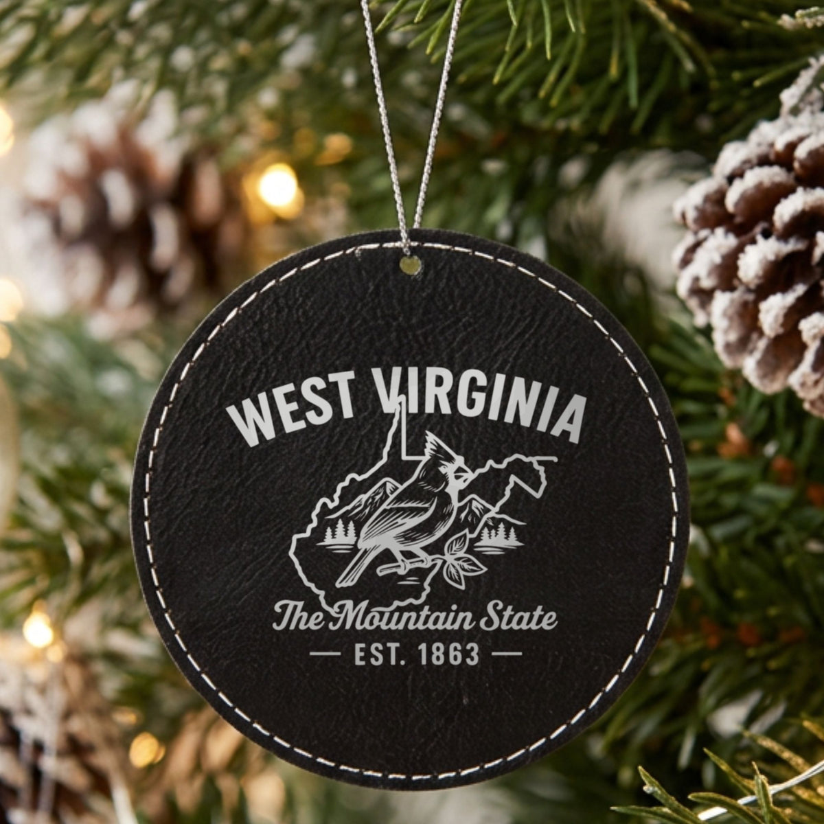 Black leatherette round Christmas ornament laser engraved with a West Virginia design of a cardinal, mountains and pines, hanging on a tree