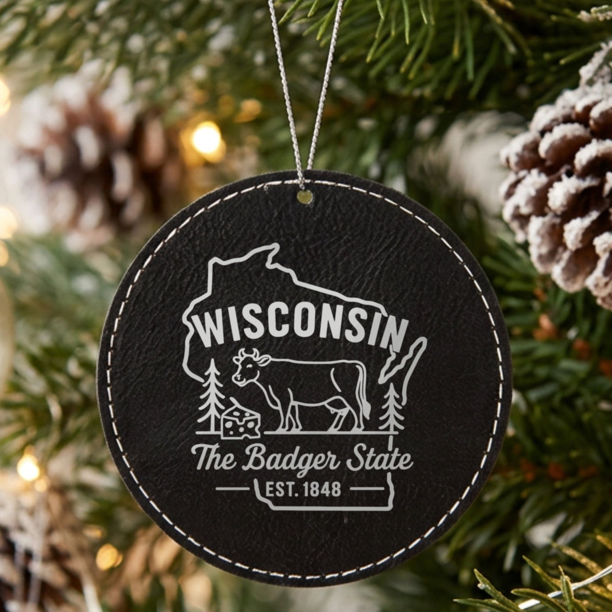 Black leatherette round Christmas ornament laser engraved with a Wisconsin design of a dairy cow and a wedge of cheese, hanging on a tree