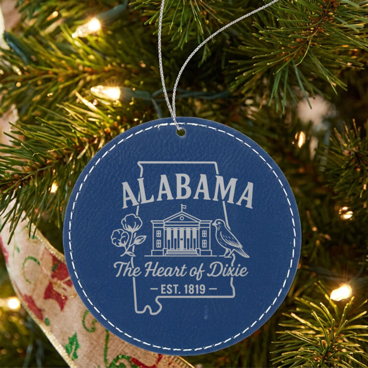 Blue leatherette round Christmas ornament laser engraved with an Alabama design of the capitol building, a cotton boll and a yellowhammer, hanging on a tree