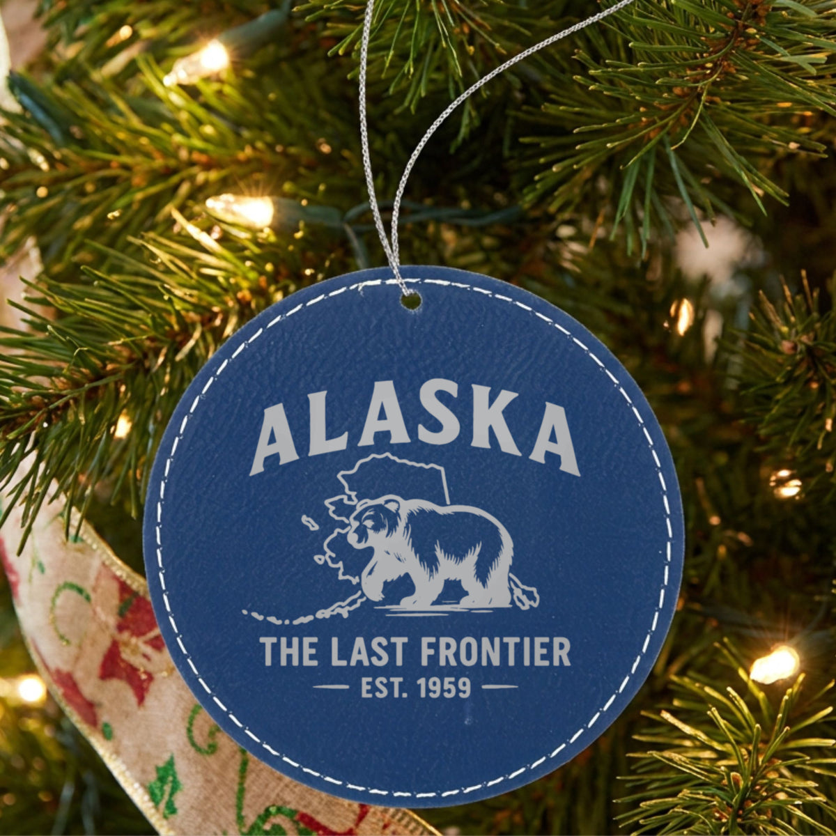 Blue leatherette round Christmas ornament laser engraved with an Alaska design of a grizzly bear crossing the state map, hanging on a tree