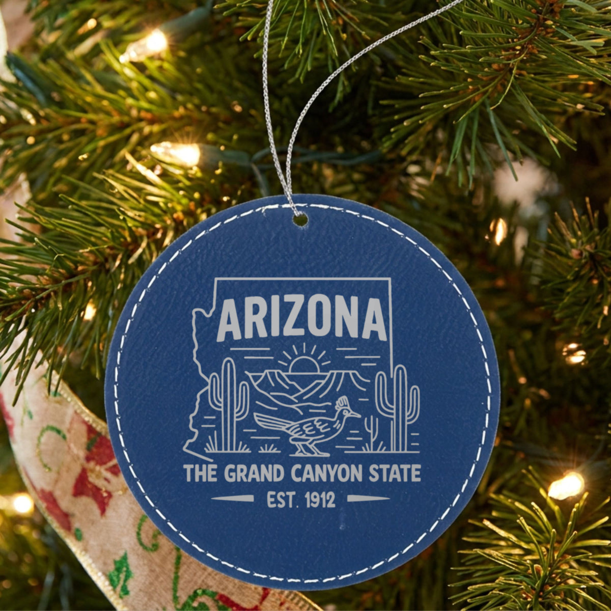 Blue leatherette round Christmas ornament laser engraved with an Arizona design of saguaro cactus, a roadrunner and the Grand Canyon at sunrise, hanging on a tree