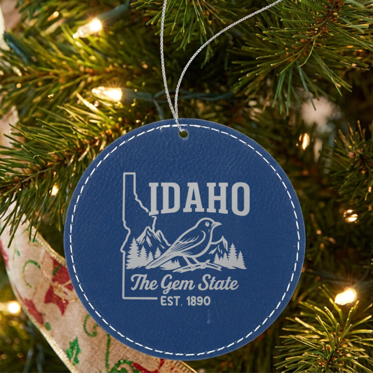 Blue leatherette round Christmas ornament laser engraved with an Idaho design of a mountain bluebird, mountains and pines, hanging on a tree