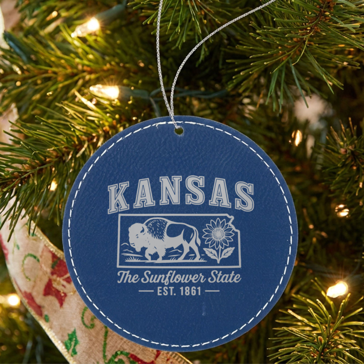 Blue leatherette round Christmas ornament laser engraved with a Kansas design of a bison and a sunflower, hanging on a tree