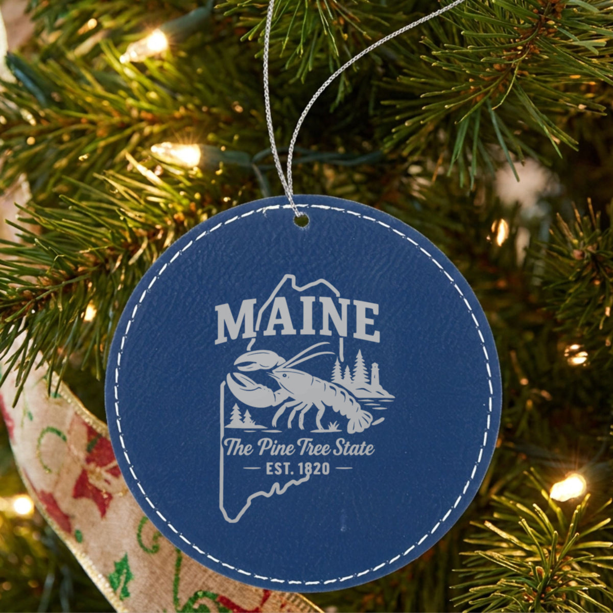 Blue leatherette round Christmas ornament laser engraved with a Maine design of a lobster, pines and a lighthouse, hanging on a tree
