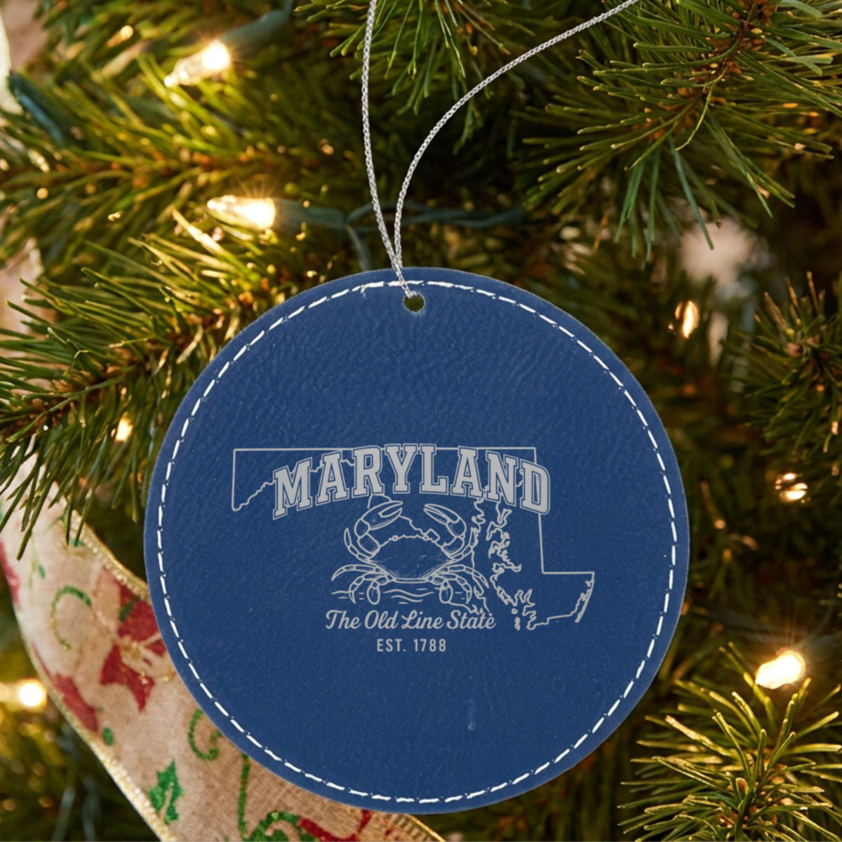 Blue leatherette round Christmas ornament laser engraved with a Maryland design of a blue crab on the Chesapeake, hanging on a tree