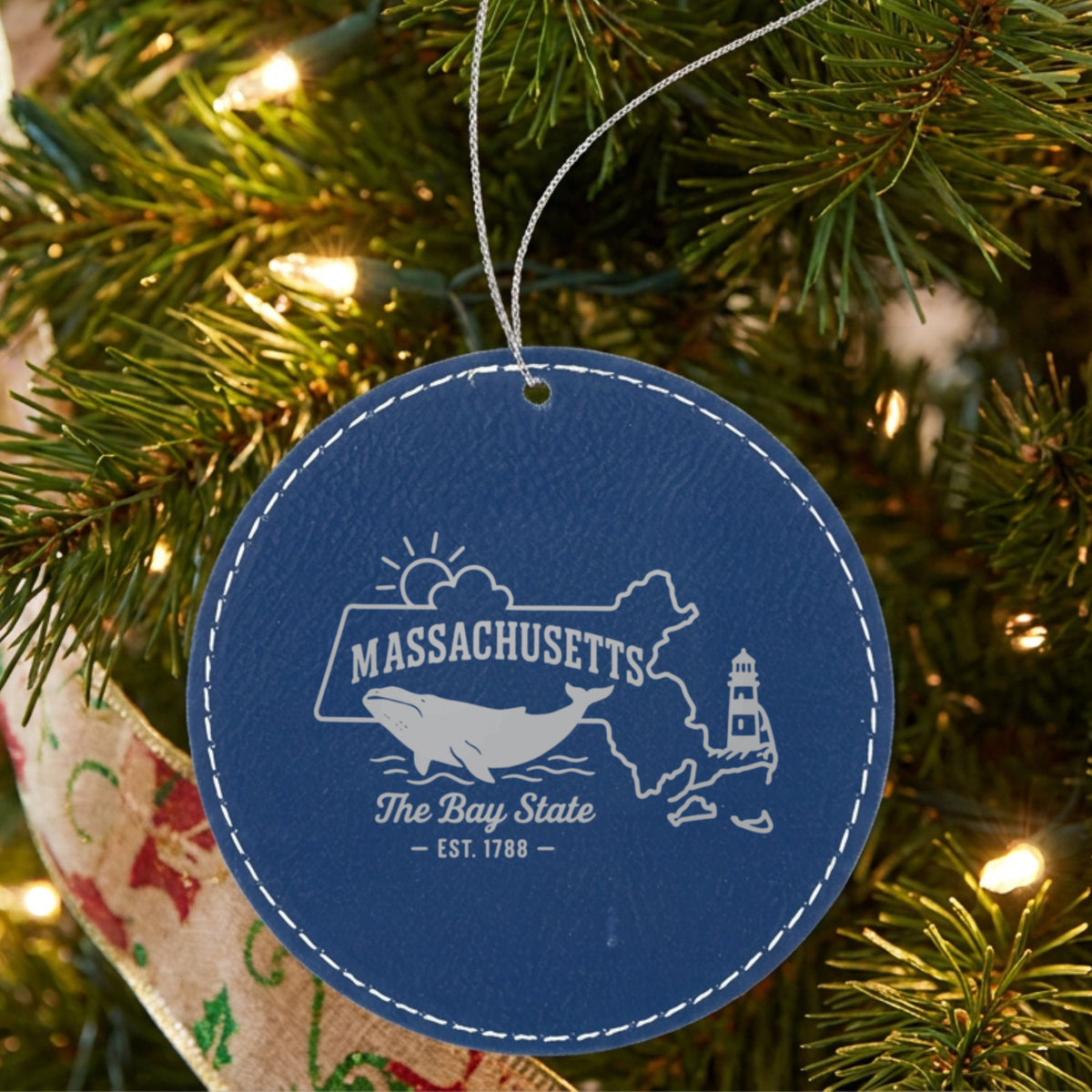 Blue leatherette round Christmas ornament laser engraved with a Massachusetts design of a whale, a lighthouse and the sun over Cape Cod and the islands, hanging on a tree