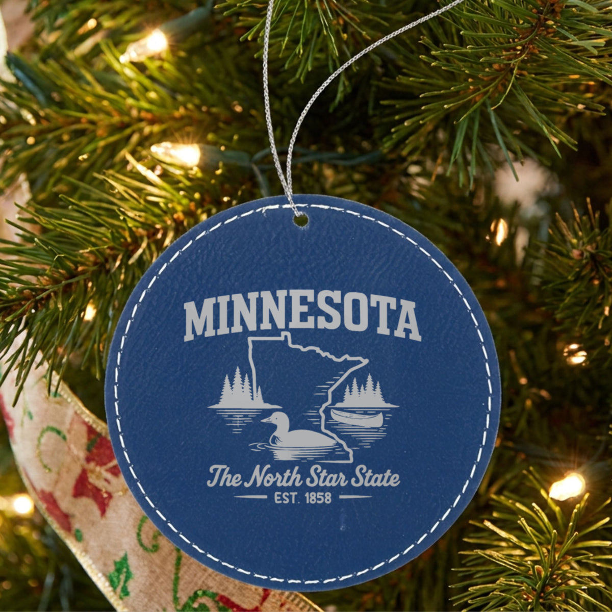 Blue leatherette round Christmas ornament laser engraved with a Minnesota design of a loon, a canoe and lakeside pines, hanging on a tree