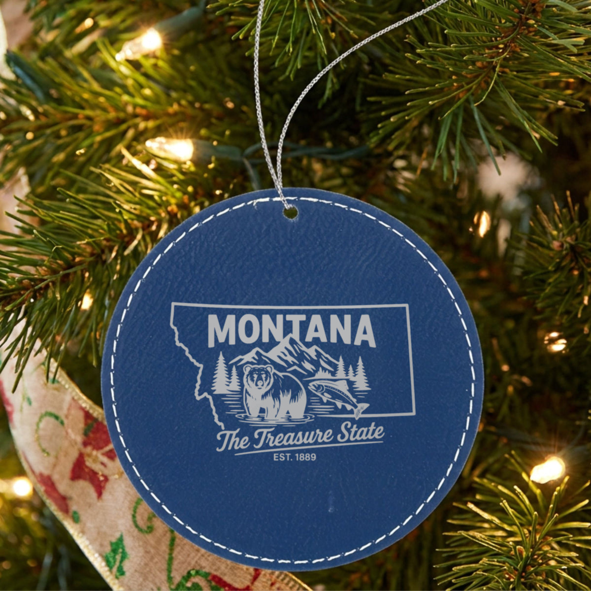 Blue leatherette round Christmas ornament laser engraved with a Montana design of a grizzly bear, a trout and mountain peaks, hanging on a tree