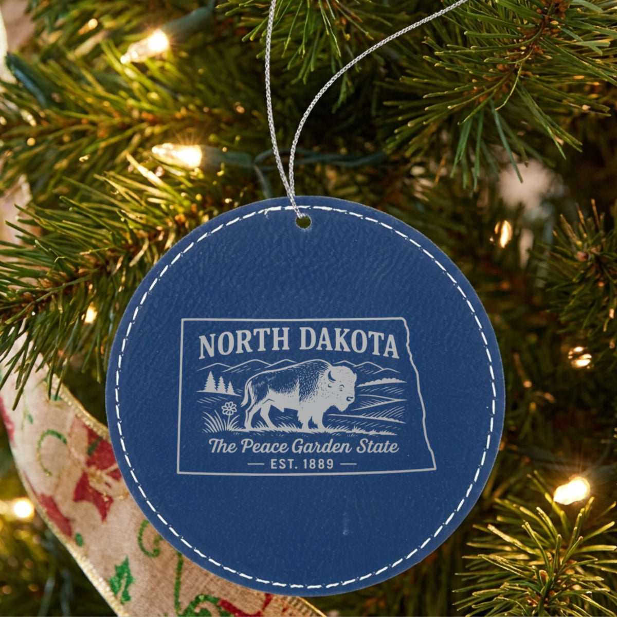 Blue leatherette round Christmas ornament laser engraved with a North Dakota design of a bison on the plains, hanging on a tree