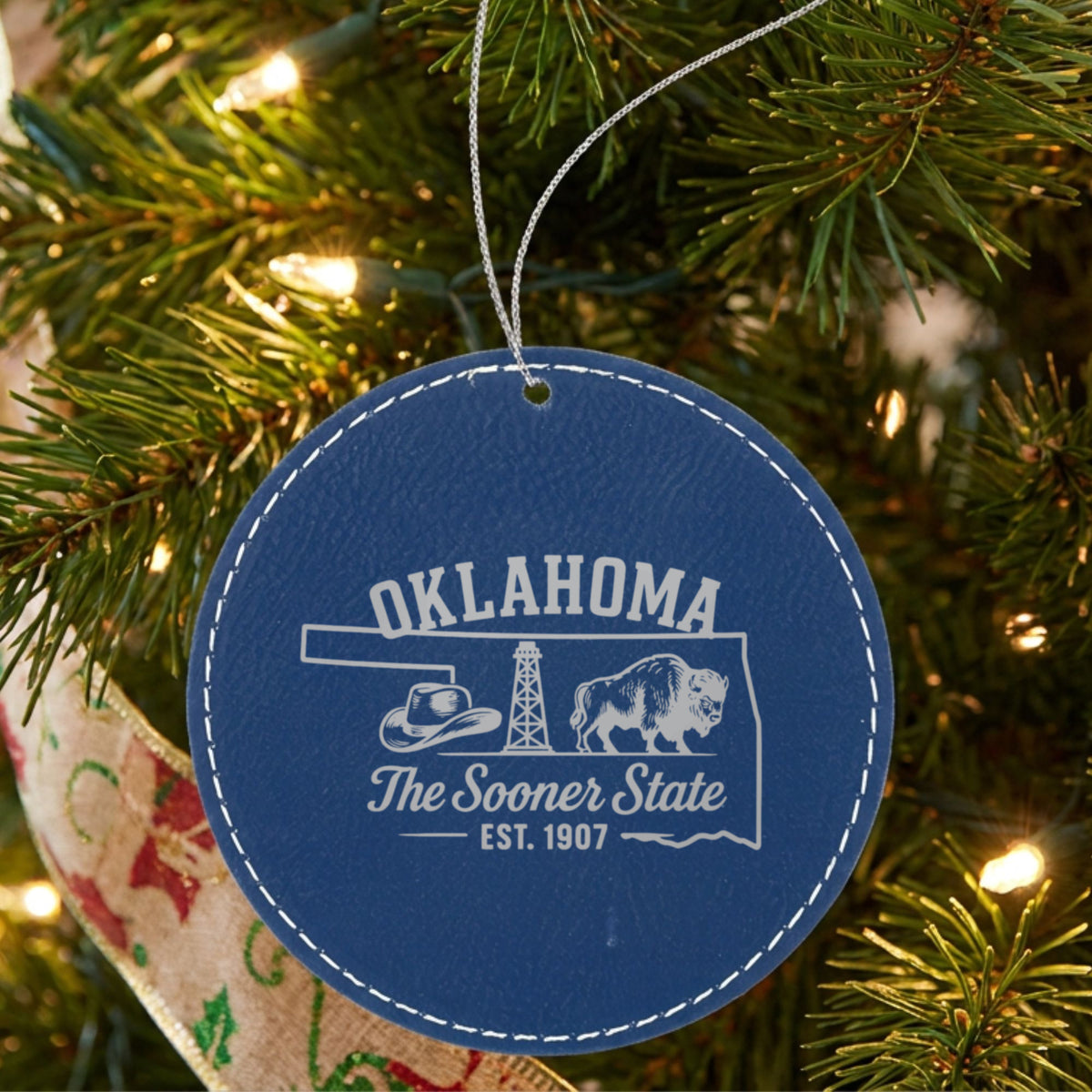 Blue leatherette round Christmas ornament laser engraved with an Oklahoma design of a cowboy hat, an oil derrick and a bison, hanging on a tree