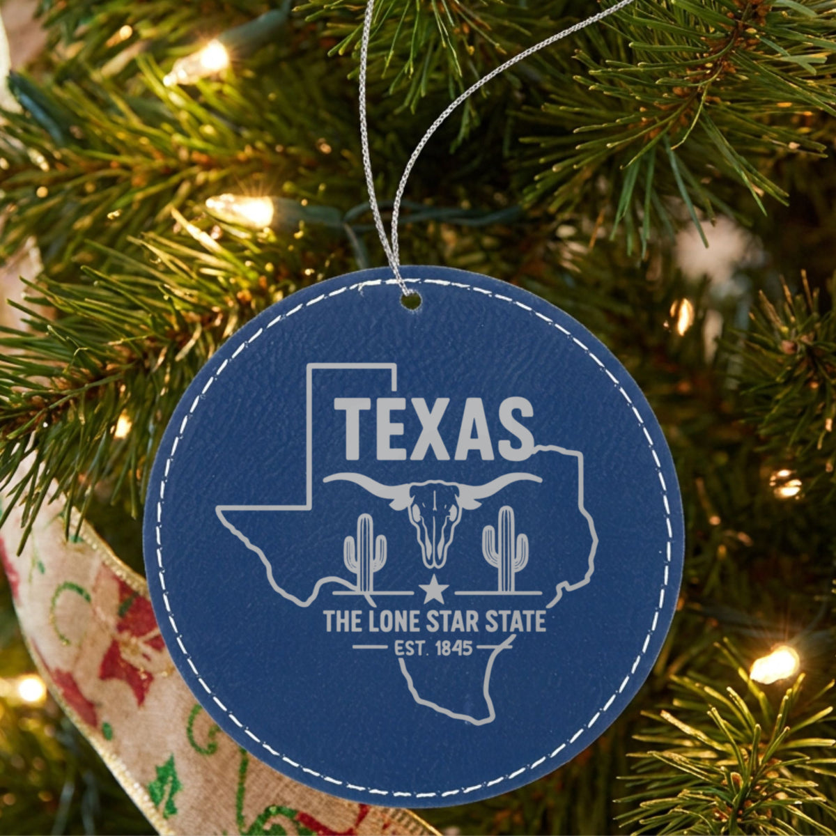 Blue leatherette round Christmas ornament laser engraved with a Texas design of a longhorn skull, cactus and a lone star, hanging on a tree