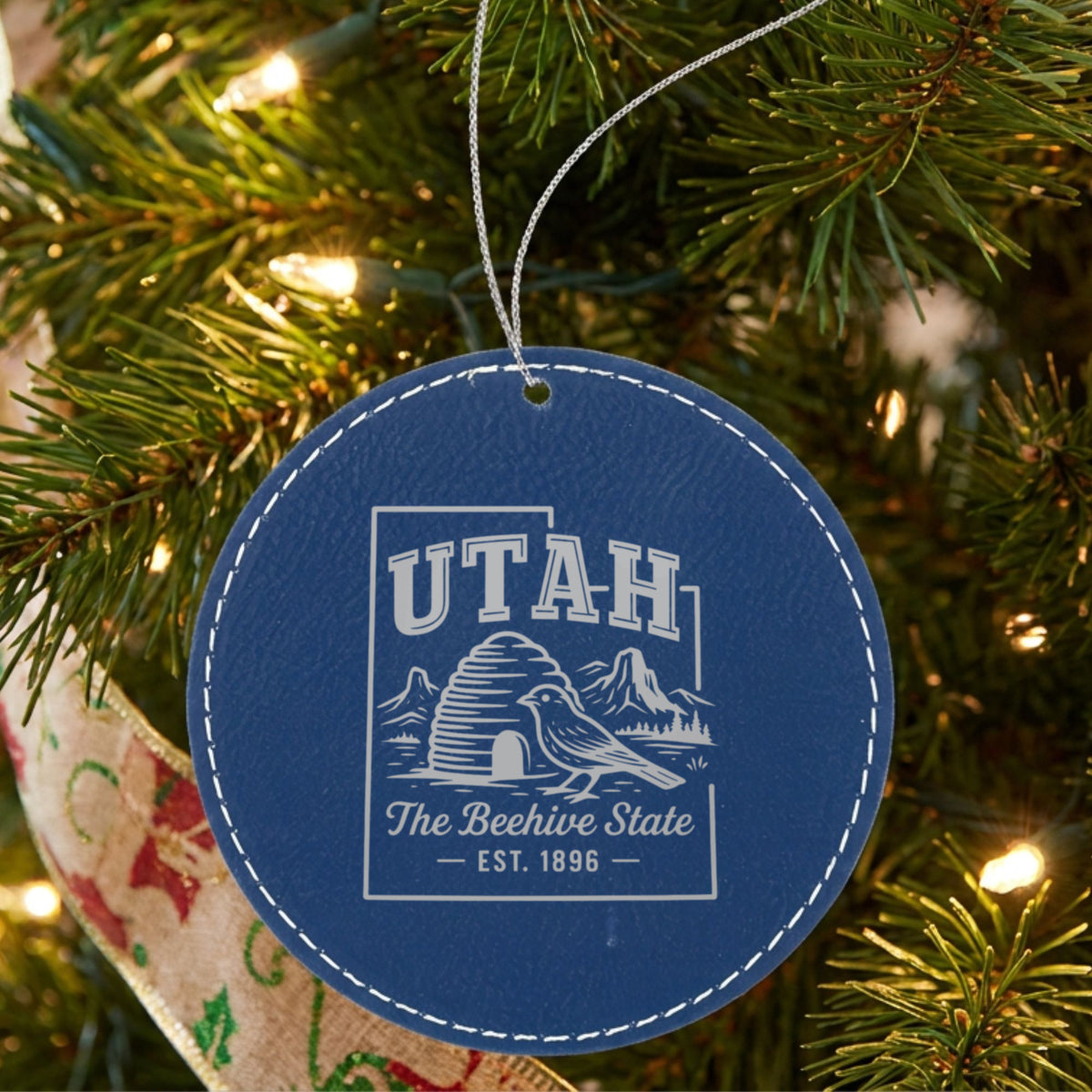 Blue leatherette round Christmas ornament laser engraved with a Utah design of a beehive, a California gull and red rock arches, hanging on a tree