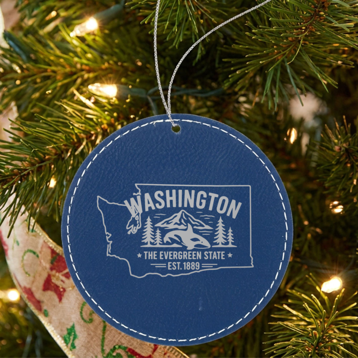 Blue leatherette round Christmas ornament laser engraved with a Washington design of an orca, Mount Rainier and evergreens, hanging on a tree