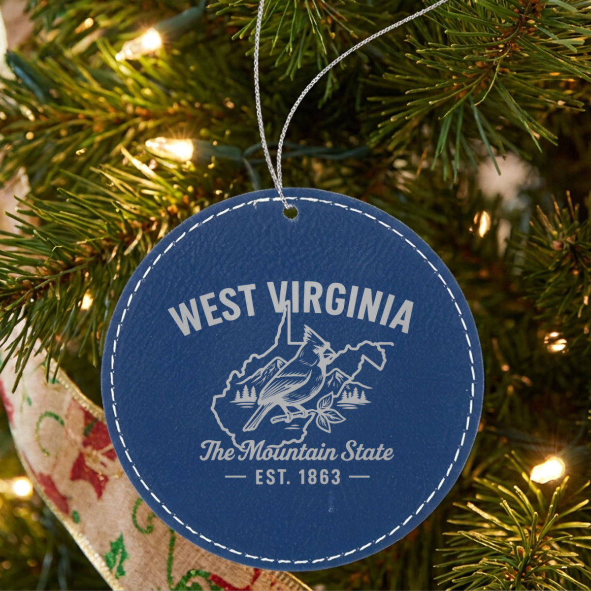 Blue leatherette round Christmas ornament laser engraved with a West Virginia design of a cardinal, mountains and pines, hanging on a tree