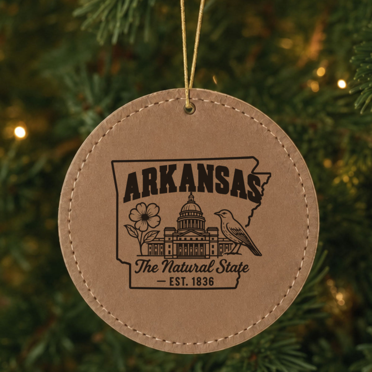 Brown leatherette round Christmas ornament laser engraved with an Arkansas design of the capitol dome, an apple blossom and a mockingbird, hanging on a tree