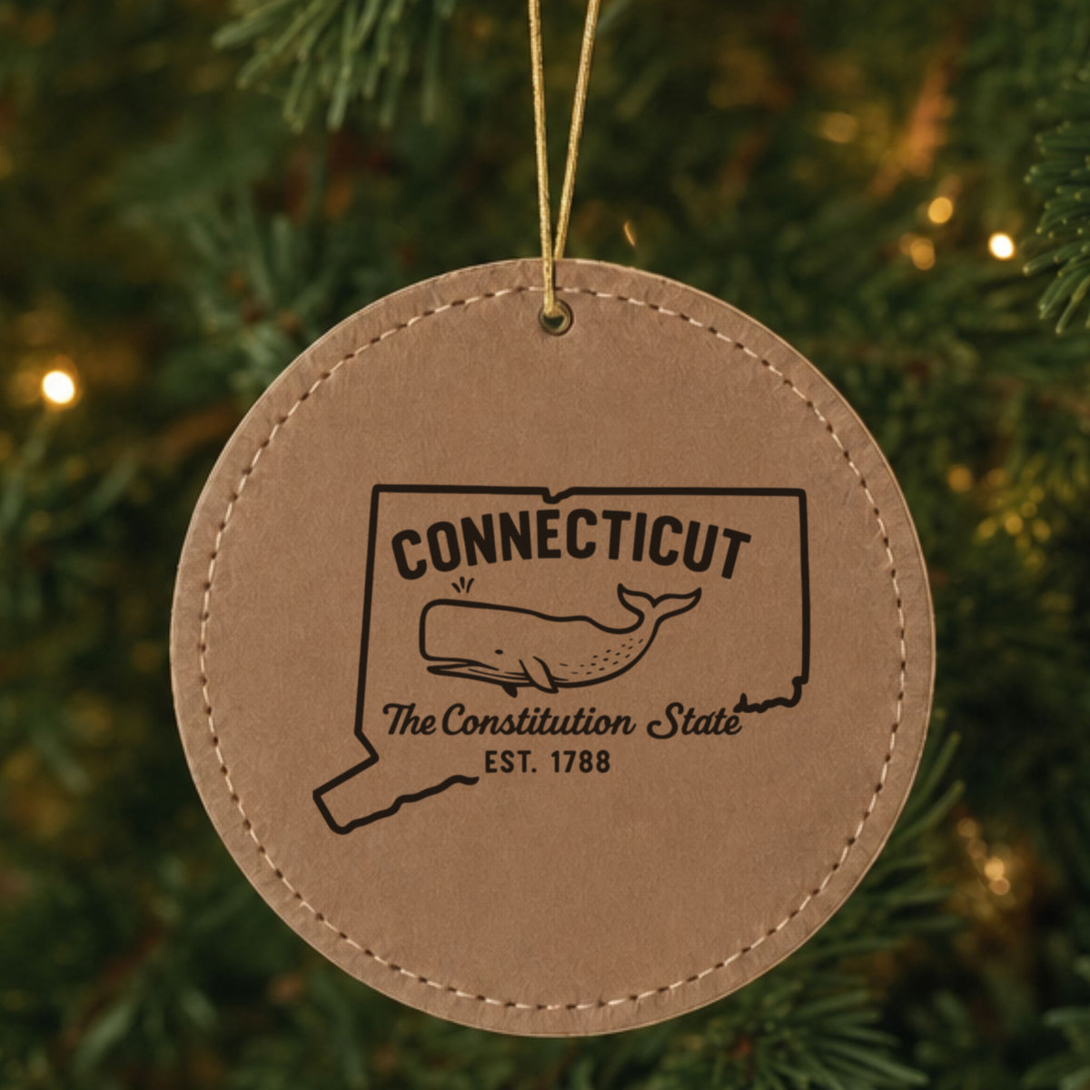 Brown leatherette round Christmas ornament laser engraved with a Connecticut design of a sperm whale, hanging on a tree