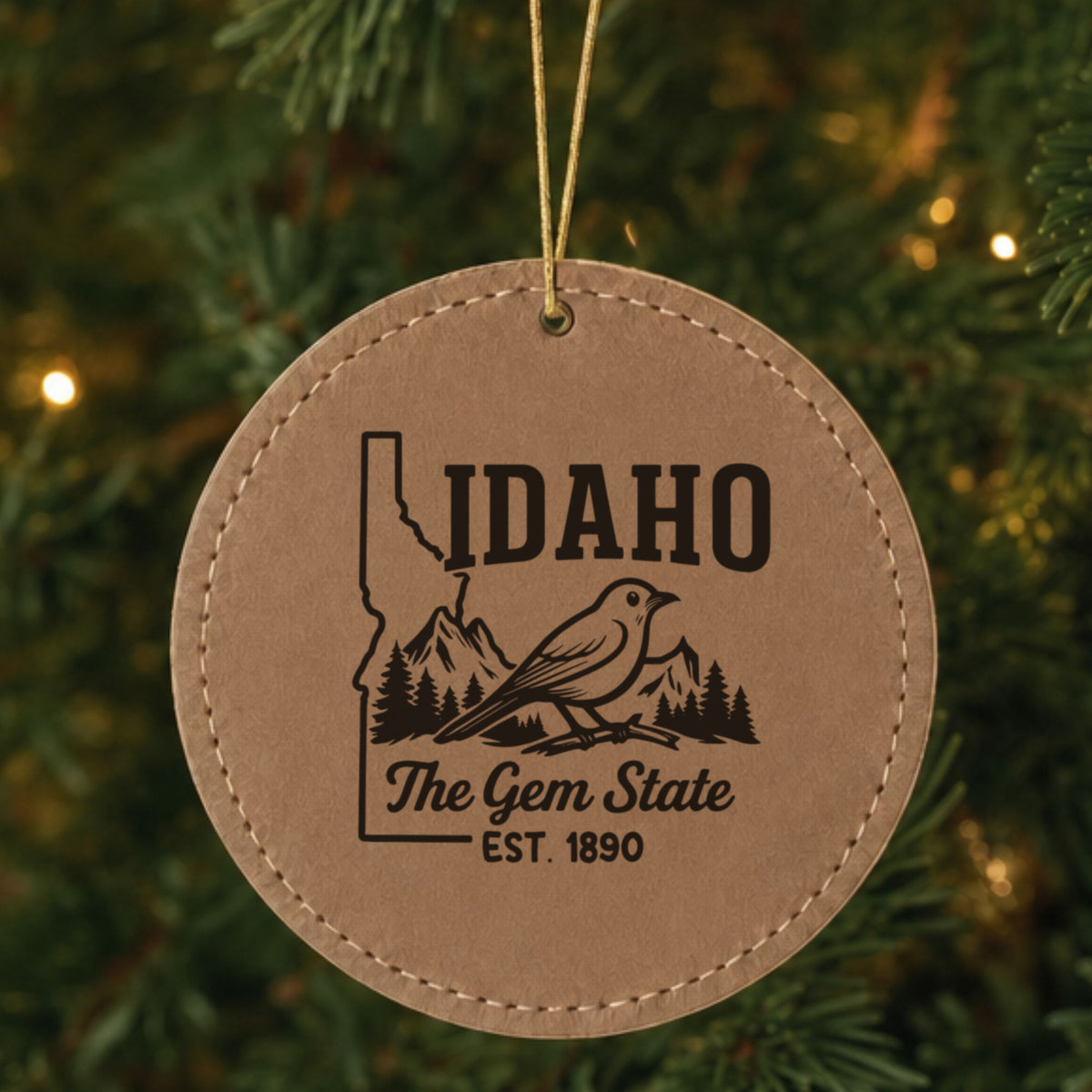 Brown leatherette round Christmas ornament laser engraved with an Idaho design of a mountain bluebird, mountains and pines, hanging on a tree