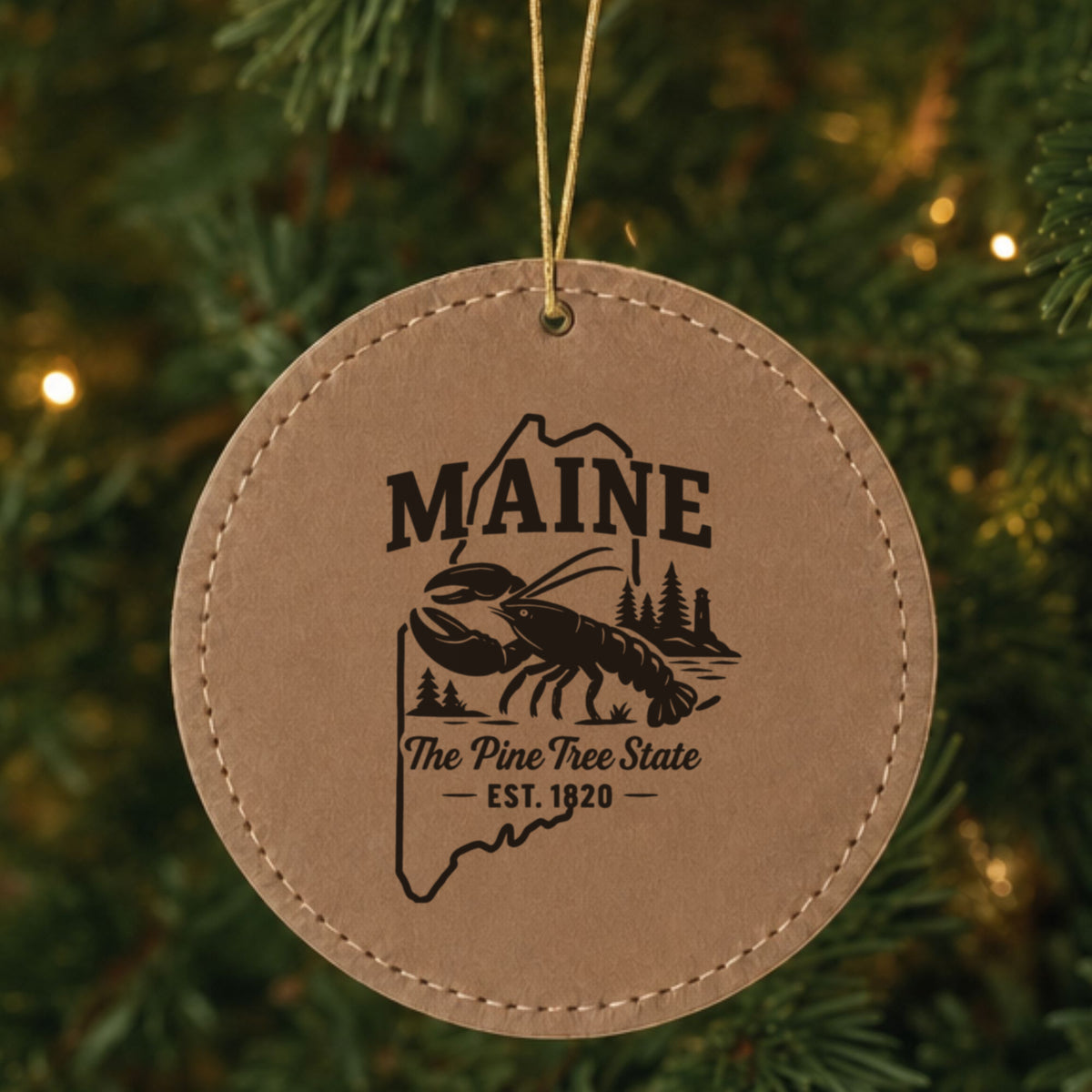 Brown leatherette round Christmas ornament laser engraved with a Maine design of a lobster, pines and a lighthouse, hanging on a tree