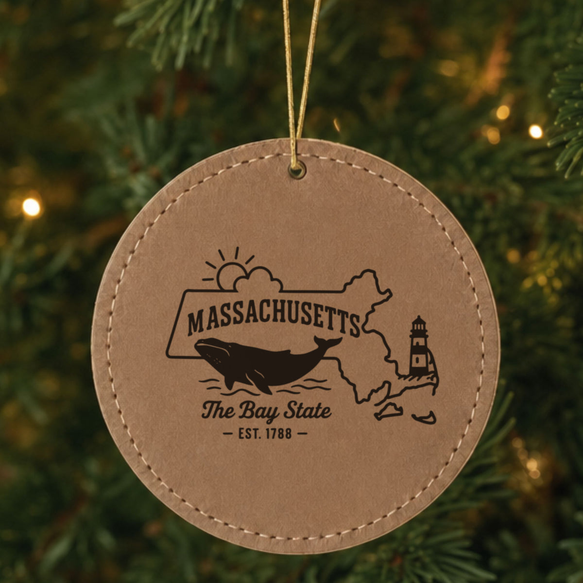 Brown leatherette round Christmas ornament laser engraved with a Massachusetts design of a whale, a lighthouse and the sun over Cape Cod and the islands, hanging on a tree