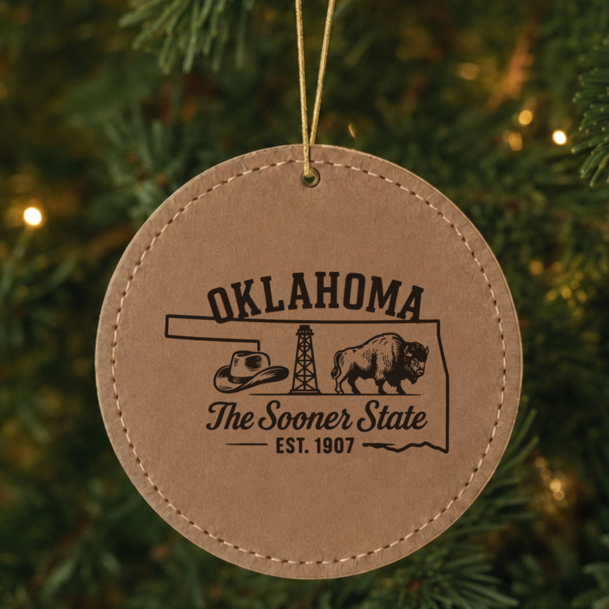 Brown leatherette round Christmas ornament laser engraved with an Oklahoma design of a cowboy hat, an oil derrick and a bison, hanging on a tree