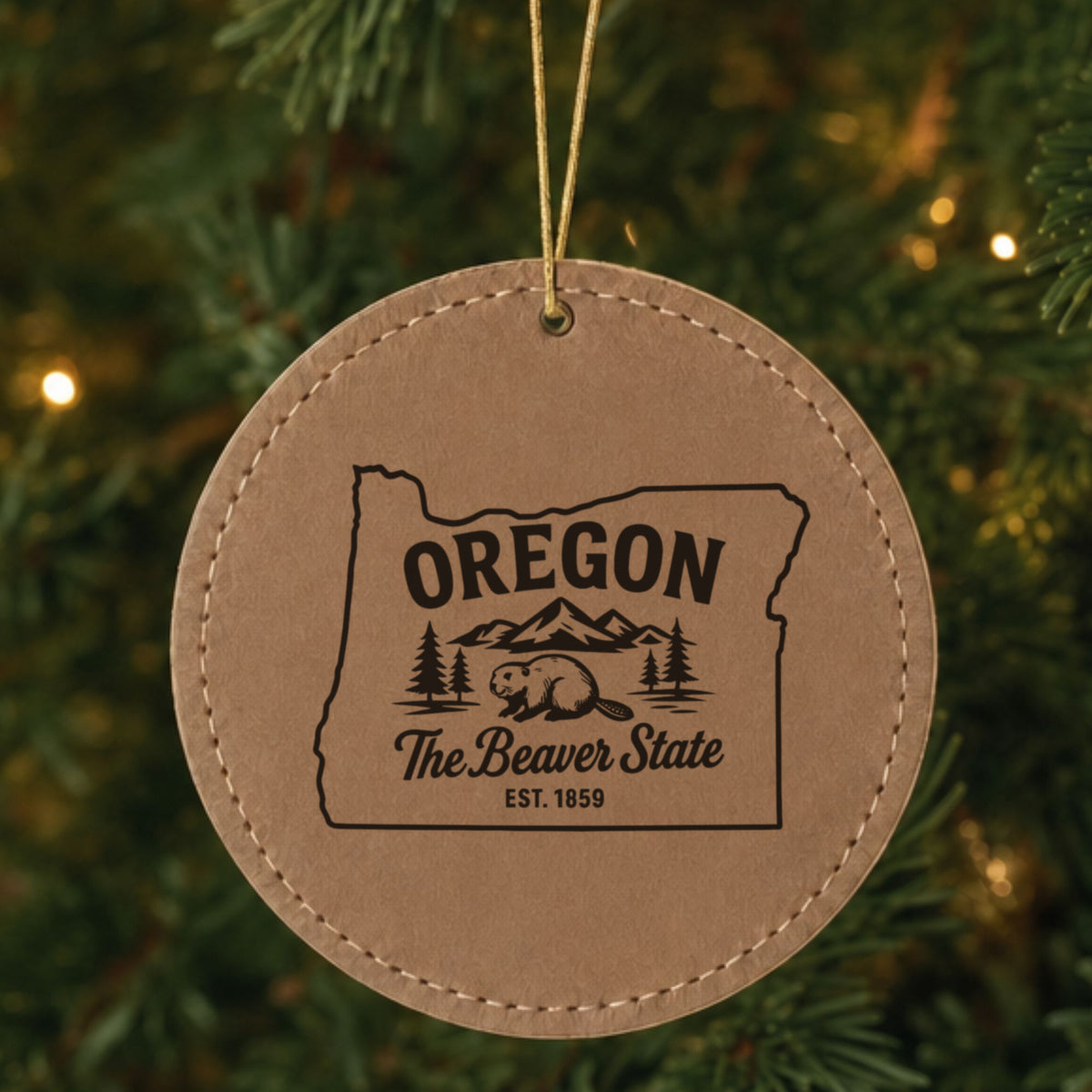 Brown leatherette round Christmas ornament laser engraved with an Oregon design of a beaver, mountains and pines, hanging on a tree