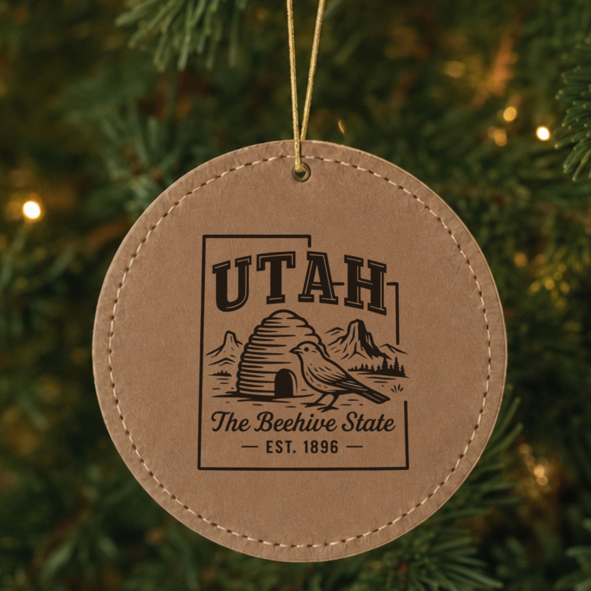 Brown leatherette round Christmas ornament laser engraved with a Utah design of a beehive, a California gull and red rock arches, hanging on a tree