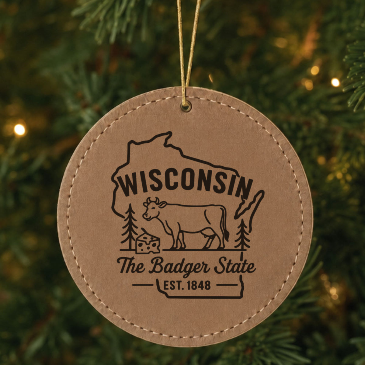Brown leatherette round Christmas ornament laser engraved with a Wisconsin design of a dairy cow and a wedge of cheese, hanging on a tree