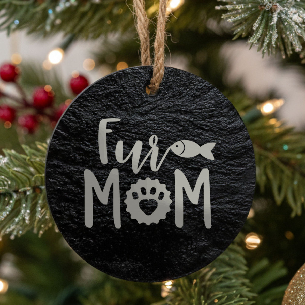 Round black slate Christmas ornament hanging from a jute string on a decorated Christmas tree, laser engraved in white with “Fur Mom Fish”.