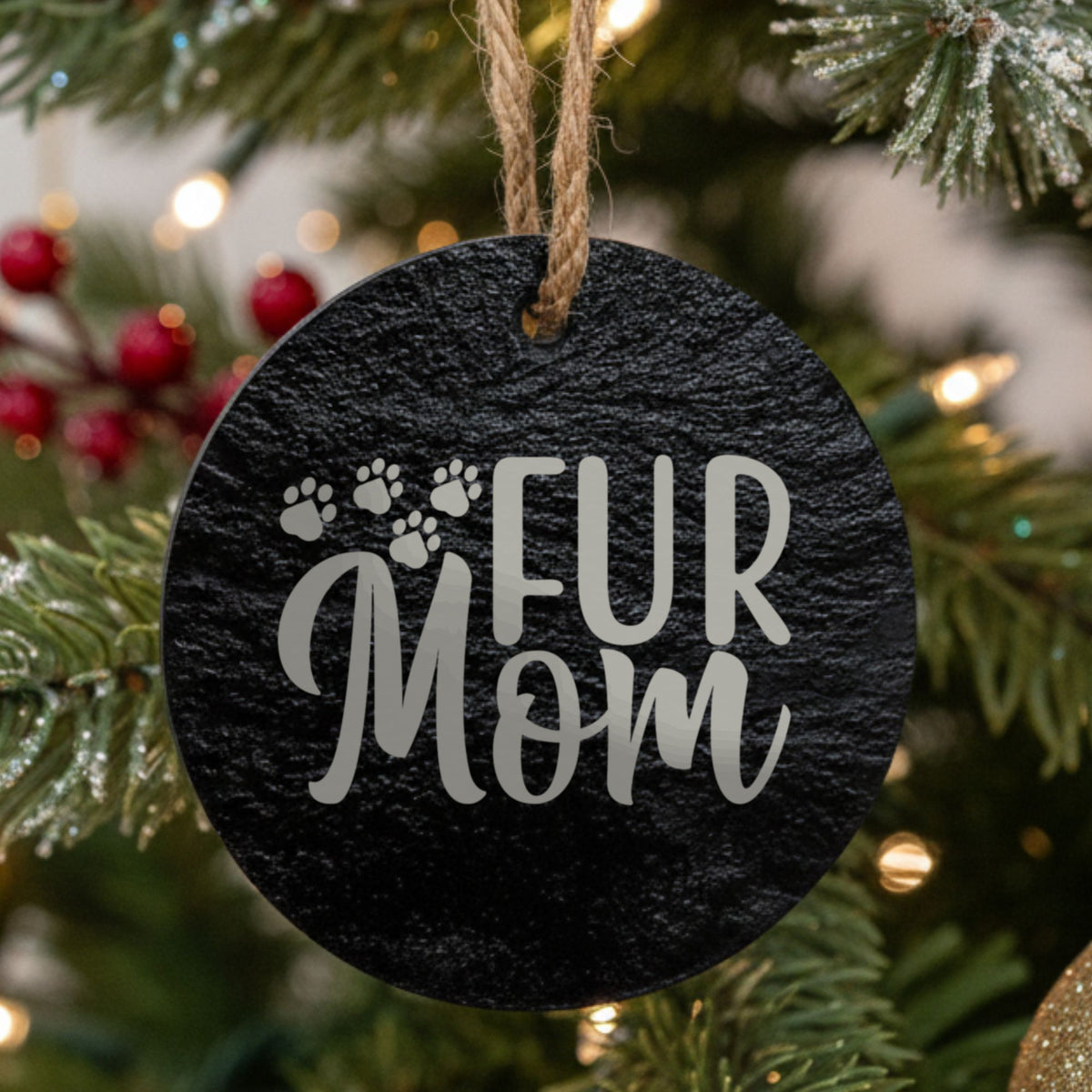Round black slate Christmas ornament hanging from a jute string on a decorated Christmas tree, laser engraved in white with “Fur Mom Paws”.