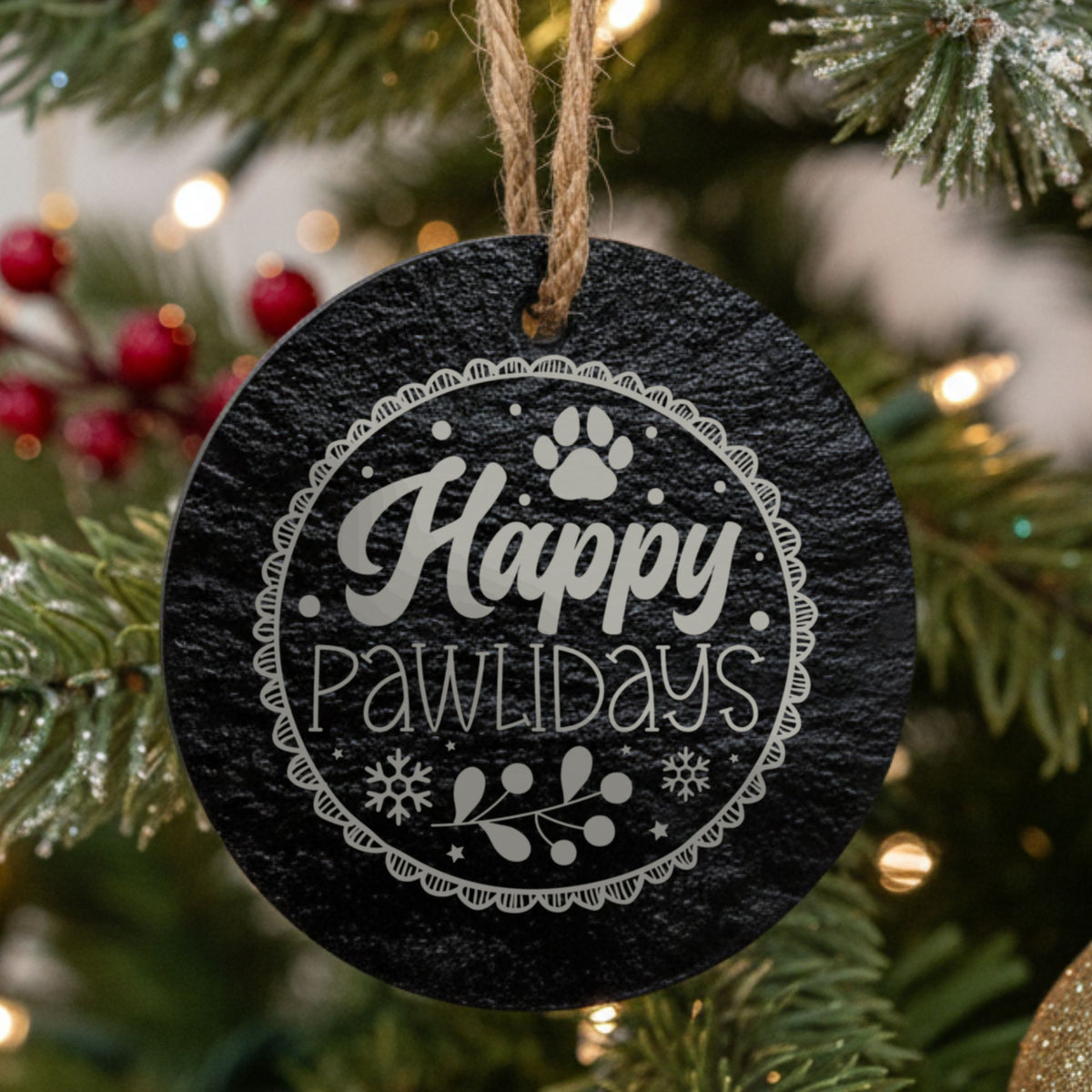 Round black slate Christmas ornament, laser engraved in white with Happy Pawlidays inside a ring of paw prints and snowflakes