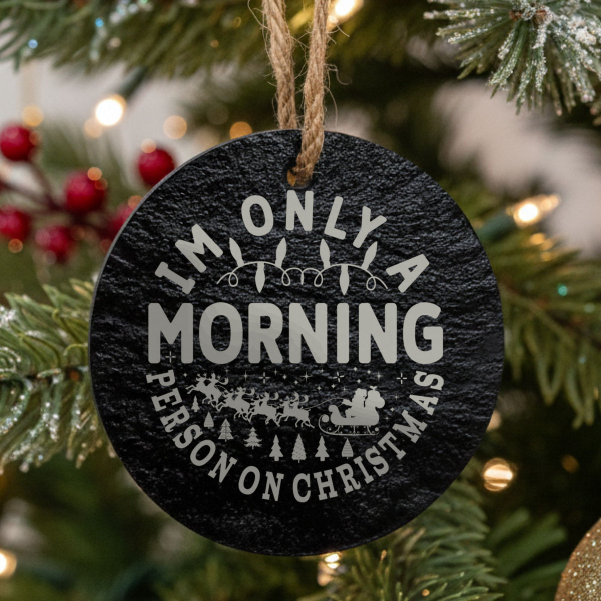 Round black slate Christmas ornament hanging from a jute string on a decorated Christmas tree, laser engraved in white with “I'm Only a Morning Person on Christmas”.