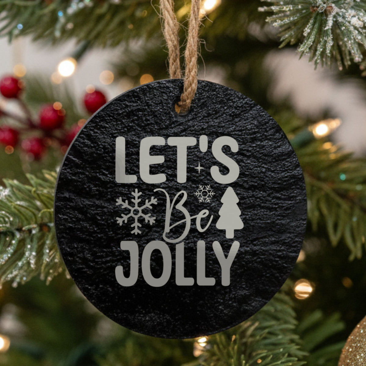 Round black slate Christmas ornament hanging from a jute string on a decorated Christmas tree, laser engraved in white with “Lets Be Jolly”.