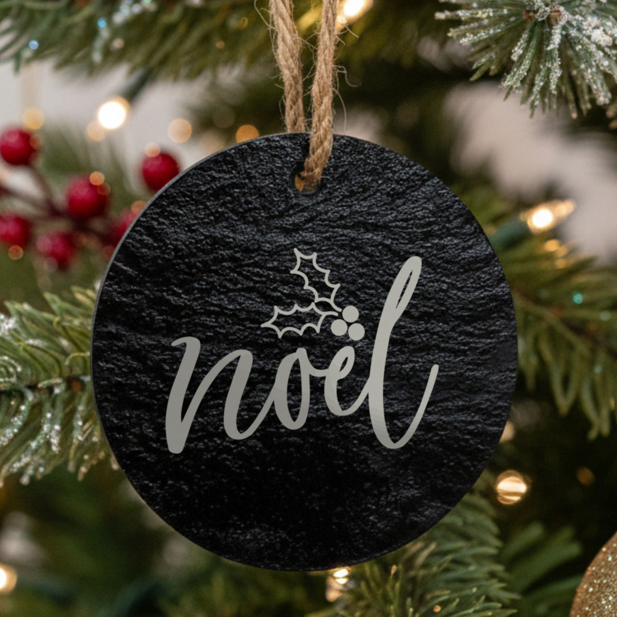 Round black slate Christmas ornament hanging from a jute string on a decorated Christmas tree, laser engraved in white with “Noel”.