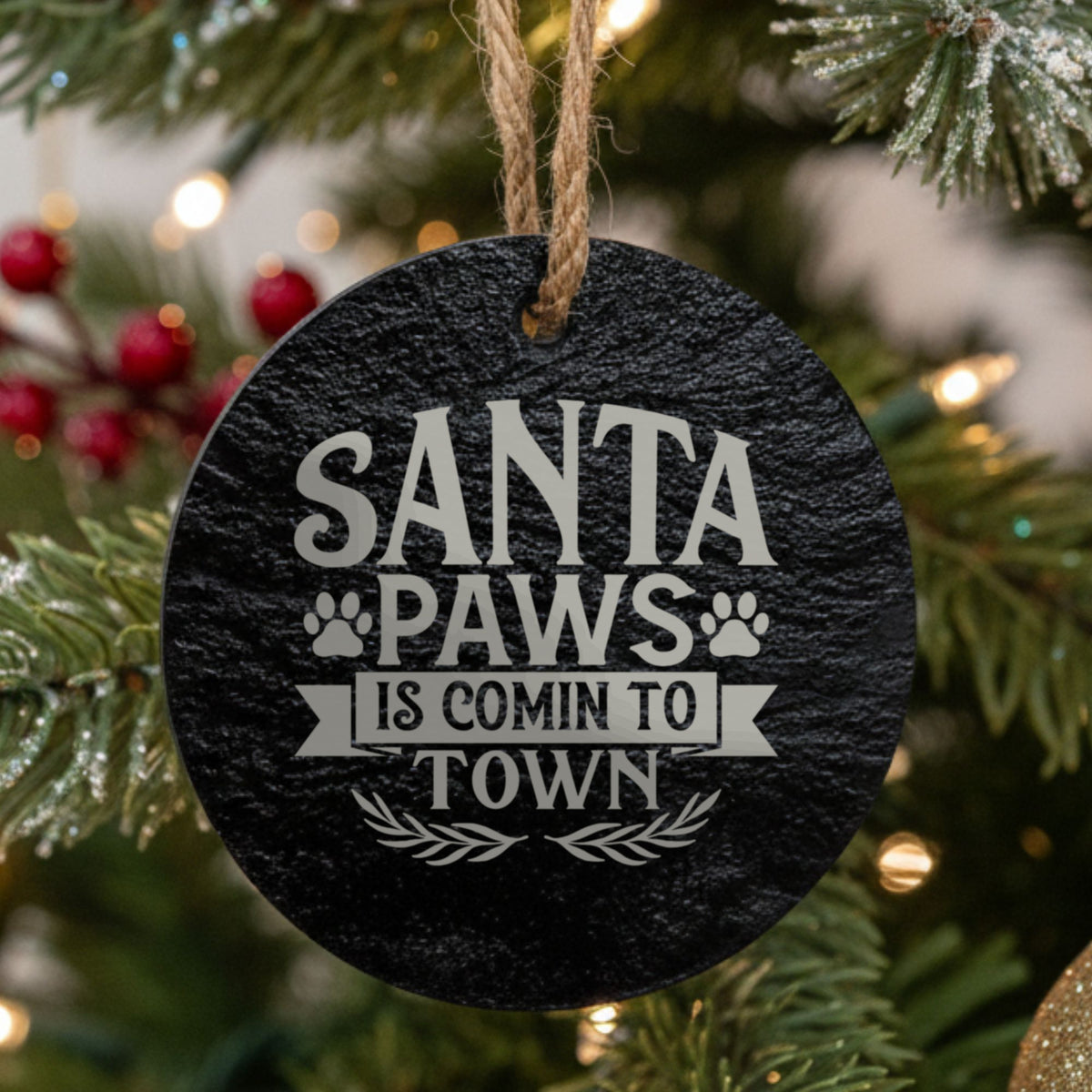 Round black slate Christmas ornament hanging from a jute string on a decorated Christmas tree, laser engraved in white with “Santa Paws is Comin to Town”.
